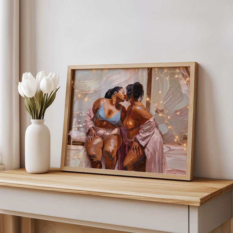 May include: A framed oil painting depicting two individuals with dark skin tones kissing. One person wears a light blue bra and pink shorts, while the other wears a pink robe. The scene is illuminated by warm string lights against a soft background.