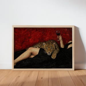 Puede incluir: Una impresión artística enmarcada que muestra a un leopardo descansando sobre una persona cubierta con tela negra sobre un fondo rojo intenso. La persona sostiene una copa de vino tinto. El leopardo tiene un pelaje manchado y mira al frente.
