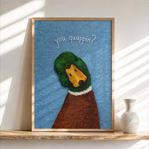 May include: A framed wall art print featuring a textured blue background with a mallard duck head and neck. The duck has a green head, a yellow bill, and a white neck ring. The white text above the duck reads, "you quappin?" in a script font.