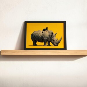 May include: A rectangular black picture frame containing a print of a grey rhinoceros standing on a solid yellow background with a small black cat resting on its back. The framed artwork is displayed on a light-colored wooden floating shelf.