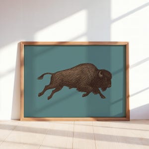 May include: A framed wall art print featuring a detailed brown ink illustration of a running American bison centered on a solid muted teal background. The artwork is enclosed in a simple light-toned wooden frame with a clean rectangular profile.