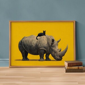 May include: A framed rectangular wall art print featuring a grey rhinoceros standing in profile against a solid yellow background. A small black cat is perched on the back of the rhinoceros. The artwork is enclosed in a light-toned wooden frame.