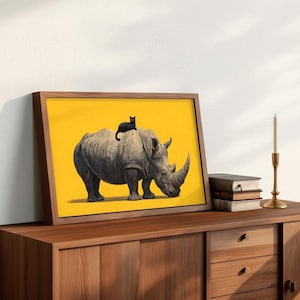 May include: A framed rectangular print featuring a gray rhinoceros standing on a solid yellow background with a small black cat perched on its back. The artwork is displayed in a wooden frame, positioned on a wooden surface next to books and a candle.