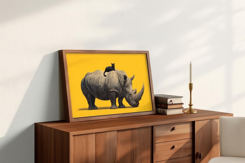May include: A framed rectangular print featuring a gray rhinoceros standing on a solid yellow background with a small black cat perched on its back. The artwork is displayed in a wooden frame, positioned on a wooden surface next to books and a candle.