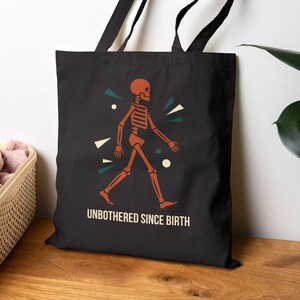 May include: Black tote bag featuring an orange skeleton graphic walking to the right, with teal and white geometric shapes. The text "UNBOTHERED SINCE BIRTH" is printed below the skeleton in white. The bag has black handles.