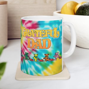 May include: White ceramic mug with a tie-dye design and the text "Grateful Dad" in yellow. The mug features a row of cartoon bears riding lawnmowers.