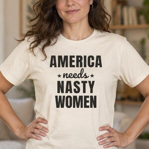 Può includere: Una t-shirt a maniche corte color crema con il testo nero "AMERICA needs NASTY WOMEN" stampato sul davanti. Il design include due piccole stelle nere ai lati della parola "needs" al centro della stampa grafica su tre righe.