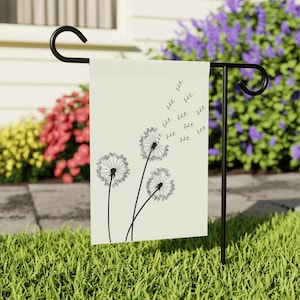 Può includere: Una bandiera da giardino color crema presenta un disegno a tratto nero di tre denti di leone con semi che volano via. La bandiera è esposta su un supporto in metallo nero in un giardino erboso, con fiori colorati sullo sfondo.