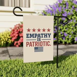 May include: A beige garden flag with the text "EMPATHY IS PATRIOTIC" in navy blue and red, with five red stars and an American flag graphic. The flag is displayed on a black metal stand in a garden setting.