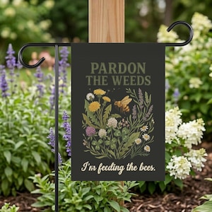 Può includere: Una bandiera da giardino nera con il testo "Pardon the Weeds" e "I'm feeding the bees." La bandiera presenta un'illustrazione colorata di fiori selvatici e un'ape. La bandiera è appesa a un supporto di metallo nero.