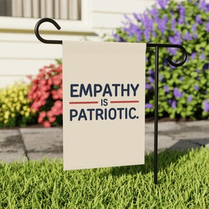 May include: A cream-colored garden flag with the words "EMPATHY IS PATRIOTIC" in navy blue text, with a red line separating the words. The flag is displayed on a black metal stand in a garden setting with colorful flowers.