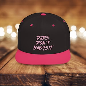 Puede incluir: Gorra de béisbol negra y rosa con el texto "DADS DON'T BABYSIT" bordado en rosa.