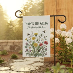 Könnte beinhalten: Eine Gartenflagge mit dem Text "Pardon the Weeds I'm feeding the bees." Die Flagge zeigt eine farbenfrohe Illustration von Wildblumen und Bienen. Die Flagge wird auf einem schwarzen Metallständer in einem Garten präsentiert.