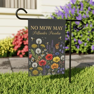 May include: A black garden flag with the text "NO MOW MAY Pollinator Paradise" in gold script. The flag features a colorful illustration of wildflowers, bees, and a butterfly. The flag is displayed on a black metal stand in a grassy yard.