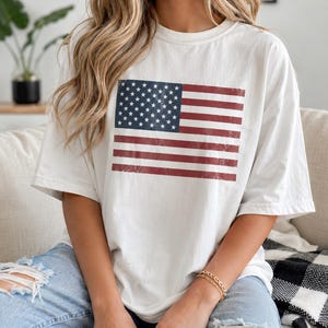 Può includere: Una t-shirt bianca a maniche corte con una grande grafica centrale della bandiera americana. Il design della bandiera presenta una texture usurata e invecchiata con i classici colori rosso, bianco e blu. La maglietta ha un girocollo e una vestibilità ampia.