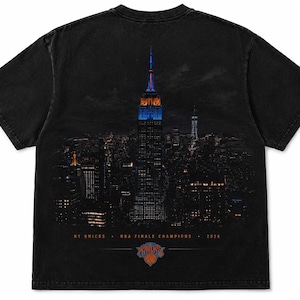 以下が含まれることがあります： 黒の半袖Tシャツ。夜のニューヨークのスカイラインが描かれており、エンパイア・ステート・ビルが青くライトアップされています。スカイラインの下には「NY KNICKS NBA FINALS CHAMPIONS 2026」という文字とチームロゴがプリントされています。