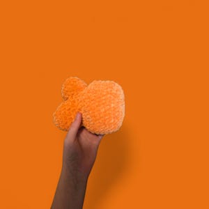 May include: A plush, orange-colored ampersand symbol held against an orange background. The ampersand is made of a soft, textured material, and the image is well-lit, highlighting the texture and shape of the symbol.