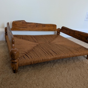 May include: A small, square, woven-seat pet bed with a wooden frame. The bed has a natural brown woven seat and a dark brown wooden frame with rounded legs and armrests. The bed is designed for a small dog or cat.