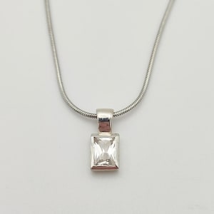 Op de afbeelding: Een zilveren ketting met een rechthoekige, heldere edelsteen hanger. De hanger is gezet in een zilveren bezel en hangt aan een fijne slangenketting. De ketting wordt getoond tegen een witte achtergrond.
