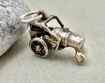 May include: A silver-coloured metal charm shaped like a vintage steamroller, featuring a prominent front roller, a central engine block, a rear driver cabin, and a circular jump ring attached to the top for hanging from a bracelet or necklace chain.