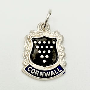 Könnte beinhalten: Ein silberner Anhänger mit einem Schilddesign. Das Schild ist schwarz mit weißen Punkten und wird von einem dekorativen Rahmen umgeben. Das Wort "CORNWALL" ist auf einem blauen Banner unten zu sehen.
