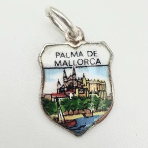 May include: A silver-toned charm with a shield shape, featuring the text "PALMA DE MALLORCA" above a colorful illustration of a coastal city with a cathedral, trees, and a sailboat. The charm has a small loop for attachment.