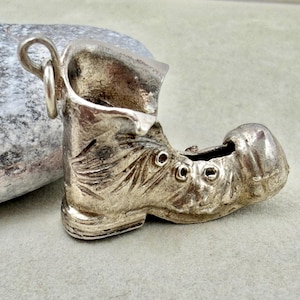 May include: A silver-colored metal charm shaped like a small boot. The charm features a textured surface with molded details including eyelets for laces and a rounded toe. A small circular jump ring is attached to the top back of the boot.
