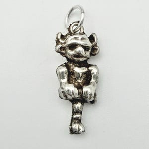 Puede incluir: Un dije plateado con forma de duende o diablillo sentado. El dije tiene una cara detallada con una nariz prominente y cabello rizado. Está unido a un pequeño anillo plateado para colgar.