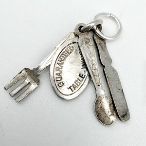 Op de afbeelding: Een zilverkleurige metalen bedel bestaande uit een miniatuurvork, een lepel, een mes en een ovaal label met de tekst GUARANTEED TABLE-WARE, bevestigd aan een ronde ring. De metalen onderdelen hebben een verweerde, doffe afwerking met zichtbare textuur.