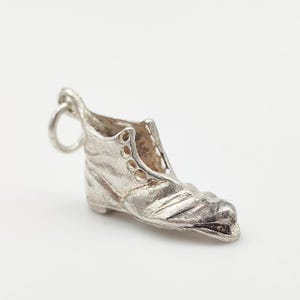 Op de afbeelding: Een kleine, zilverkleurige laarshanger met een lus om aan een armband of ketting te bevestigen. De hanger heeft gedetailleerde stiksels en oogjes en is waarschijnlijk gemaakt van sterling zilver. De laars is een miniatuurreplica van een voetbalschoen.