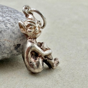 May include: A small, silver-toned metal charm shaped like a seated creature with pointed ears and a mischievous facial expression. The figure is depicted in a hunched, sitting position with its arms wrapped around its knees, featuring a jump ring.