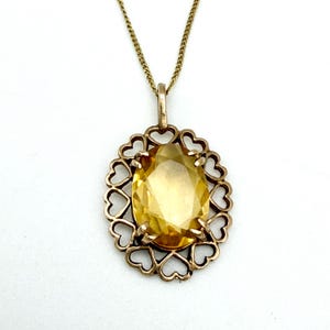 May include: A gold pendant necklace with a large, oval yellow gemstone. The gemstone is set within a heart-shaped filigree frame. The necklace chain is gold, creating an elegant piece of jewellery. The pendant is a striking design.