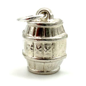 May include: A silver-toned metal charm shaped like a small barrel. The barrel features vertical staves, horizontal bands, and a series of small, embossed cross-like symbols around the center. A circular jump ring is attached to the top loop.