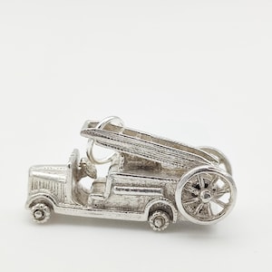 Op de afbeelding: Een zilverkleurige miniatuur brandweerwagenbedel met een ladder en gedetailleerde wielen. De bedel heeft een vintage uitstraling en is waarschijnlijk gemaakt van metaal. Het ontwerp omvat een cabine en een omhooggaande ladder.