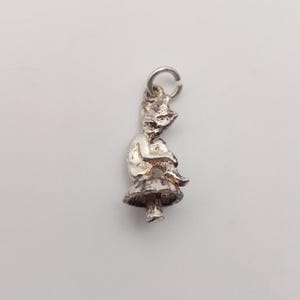May include: A silver charm of a fairy seated on a mushroom. The fairy is depicted with its arms around its knees. The mushroom has a detailed cap and stem. The charm has a loop for attaching to a bracelet or necklace. The charm is made of silver.