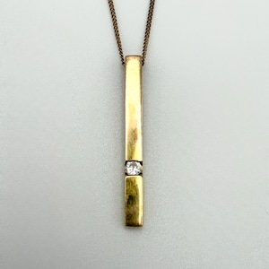 May include: A gold-coloured pendant necklace with a rectangular bar design. The bar features a small, clear gemstone set in the centre. The necklace chain is a matching gold colour. The pendant hangs vertically.