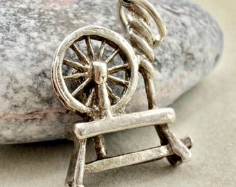 May include: A silver-toned metal charm shaped like a spinning wheel, featuring a circular wheel with spokes, a central support post, and a horizontal base. The charm includes a small circular jump ring attached to the top for hanging purposes.