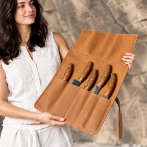 Peut inclure: Un étui à couteaux en cuir beige doté de quatre compartiments, chacun contenant un couteau de cuisine avec un manche en bois et une lame en métal. L'étui possède un rabat supérieur et une longue lanière latérale pour le maintenir fermé.