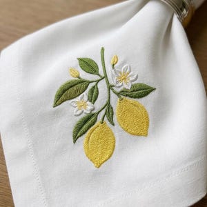 May include: White linen napkin with embroidered lemon branch design. The embroidery features yellow lemons, green leaves, and white flowers. A silver napkin ring holds the napkin in place.