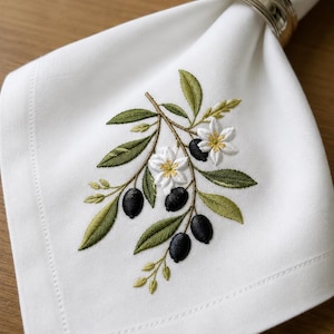 May include: White linen napkin with an embroidered olive branch design. The embroidery features green leaves, black olives, and white flowers with yellow centers. A silver napkin ring holds the napkin.