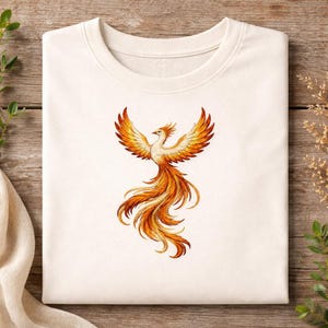 Könnte beinhalten: Cremefarbenes Sweatshirt mit einem Phönix-Design. Der Phönix hat ausgebreitete Flügel und fließende Schwanzfedern in Orange, Gelb und Gold. Das Kunstwerk ist mittig auf der Vorderseite des gefalteten Kleidungsstücks angeordnet.