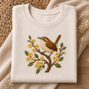 Könnte beinhalten: Cremefarbenes Sweatshirt mit einem gestickten Vogel auf einem blühenden Ast. Der Vogel hat braune und beige Federn. Gelbe Blüten und grüne Blätter schmücken den Ast und schaffen ein von der Natur inspiriertes Design. Das Sweatshirt ist gefaltet.