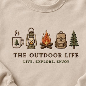 May include: Beige sweatshirt with embroidered outdoor-themed graphics. Features a mug, lantern, campfire, backpack, and pine tree. Text reads "THE OUTDOOR LIFE" and "LIVE. EXPLORE. ENJOY."