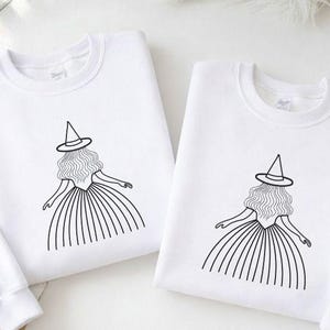 May include: Two white sweatshirts with a black line drawing of a witch wearing a hat and dress. The witch's hair is wavy, and her arms are outstretched. The sweatshirts are on a white surface.