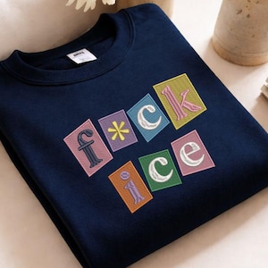 May include: Navy blue sweatshirt with the word "F*ck Ice" embroidered in colorful, block letters. The letters are outlined in pink, white, and green. The sweatshirt is made of a soft material.