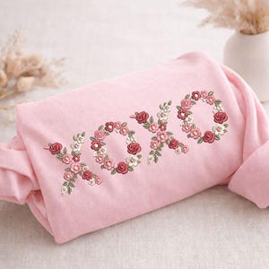 May include: A light pink sweatshirt with the embroidered letters "XOXO" in a floral design. The flowers are in shades of pink, red, and white, with green leaves. The sweatshirt is folded, with the sleeves visible.