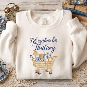 Puede incluir: Sudadera color crema con la frase "I'd rather be Thrifting" en escritura azul marino. Un gráfico de carrito de compras está lleno de porcelana azul y blanca, una lámpara y una taza de té. La sudadera tiene mangas largas.