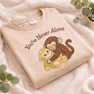 May include: Beige sweatshirt with the embroidered text "You're Never Alone." The design features two hugging cartoon monkeys, one brown and one gold. The sweatshirt is folded on a soft, textured surface.
