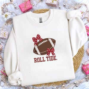 Könnte beinhalten: Weißes Sweatshirt mit brauner Football-Grafik und roten Tupfen-Schleifen. Der Text "ROLL TIDE" ist rot aufgedruckt. Das Sweatshirt hat lange Ärmel und einen Rundhalsausschnitt.