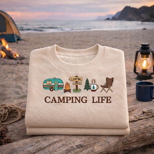 May include: Beige sweatshirt with the words "CAMPING LIFE" and camping-themed graphics. The design includes a teal camper, campfire, coffee sign, pine tree, coffee cup, and a camping chair. The sweatshirt is folded on a wooden surface.